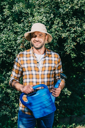 handsome male farmer in checkered shirt holding watering can and smiling at camera outdoorsの写真素材