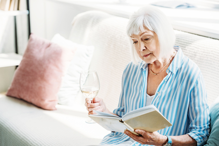 focused senior woman with glass of wine reading book on couch at homeの写真素材