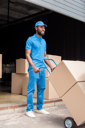 smiling african american delivery man holding cart with boxes and looking awayの写真素材