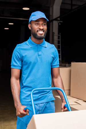 smiling african american delivery man holding cart with boxesの写真素材