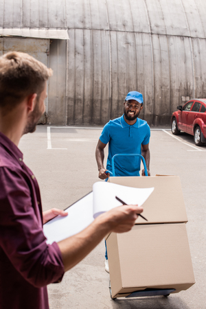 smiling african american courier with cart of boxes looking at clientの写真素材