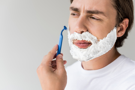 portrait of attractive man with foam on face and razor shaving beard isolated on greyの写真素材