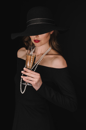 elegant woman with eyes covered by black straw holding champagne glass isolated on black backgroundの写真素材