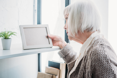 side view of senior lady looking at empty photo frame on bookshelf at homeの写真素材