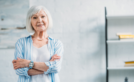 portrait of beautiful smiling senior lady in stylish clothing with arms crossed looking at camera at homeの写真素材