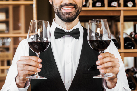 cropped shot of smiling young sommelier holding two glasses of red wine at wine storeの写真素材