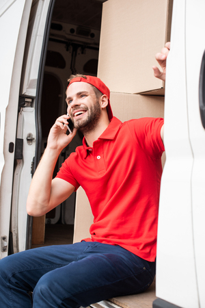 smiling delivery man in red uniform talking on smartphone while resting in vanの写真素材
