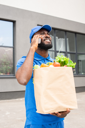 african american delivery man holding paper bag with vegetables and talking by smartphoneの写真素材