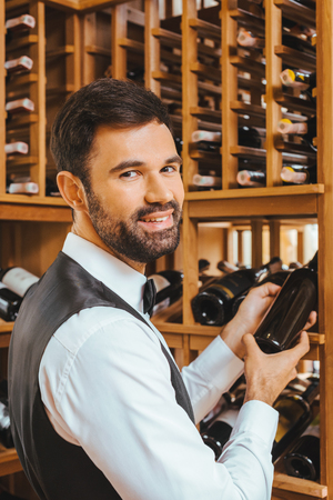 handsome young sommelier taking bottle from shelf at wine store and looking at cameraの写真素材
