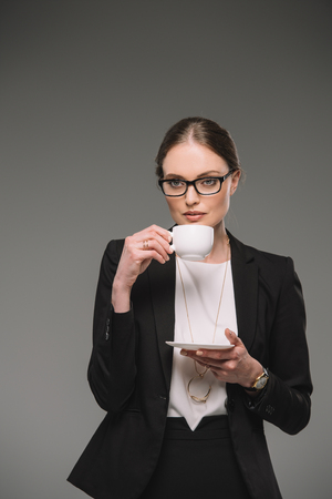 attractive businesswoman in eyeglasses drinking coffee from cup isolated on grey backgroundの写真素材