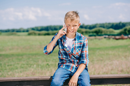 happy child talking by smartphone while sitting on fence and smiling at cameraの写真素材