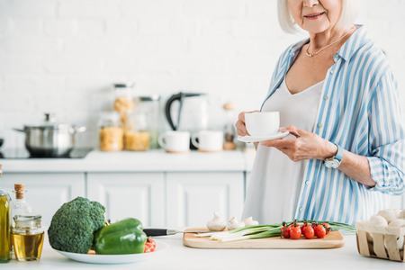cropped shot of senior woman with cup of coffee standing at counter with fresh vegetables for dinner in kitchenの写真素材