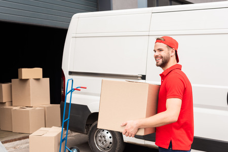 smiling delivery man in uniform with cardboard box standing near white van in street
