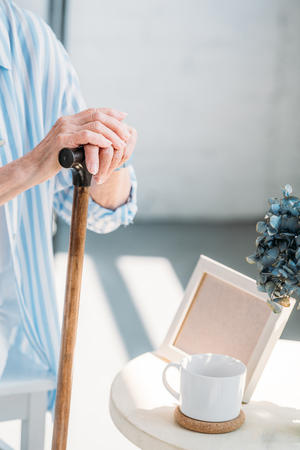 partial view of senior woman with walking stick at coffee table with cup of coffee and empty photo frameの写真素材