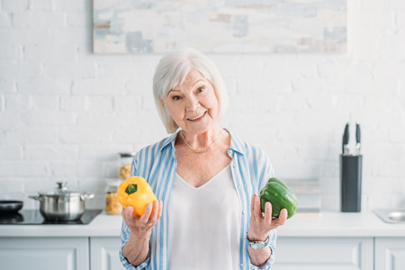 portrait of smiling senior lady with fresh bell peppers in hands in kitchenの写真素材