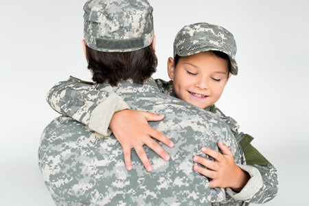 partial view of father and smiling son in military uniforms hugging each other on grey backgroundの写真素材