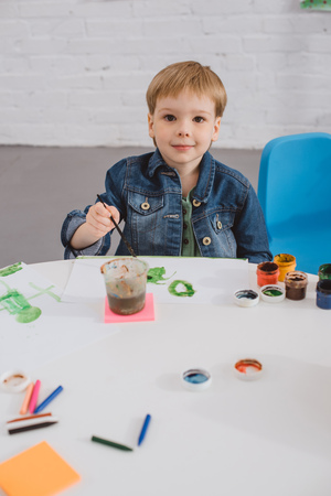 portrait of little boy sitting at table with paints and paint brush for drawing in classroomの写真素材