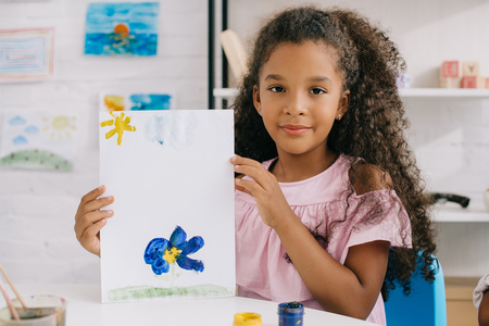portrait of african american kid showing colorful picture in hands while sitting at table in roomの写真素材