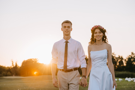 beautiful happy young wedding couple holding hands and smiling at camera while walking in park at sunsetの写真素材