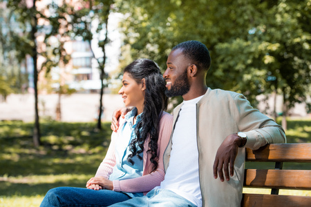 smiling african american couple sitting on wooden bench in park and looking awayの写真素材