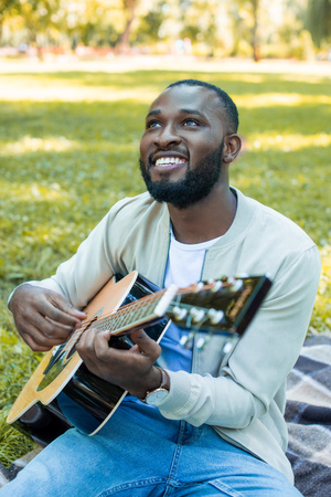 happy handsome african american man playing acoustic guitar in park and looking up