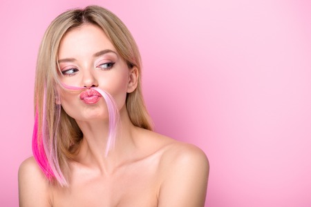 attractive young woman making mustache with pink hair strand isolated on pinkの写真素材