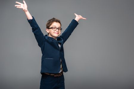schoolboy in businessman costume standing with raised hands isolated on greyの写真素材