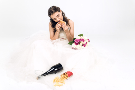 high angle view of young bride in wedding dress eating fast food while sitting on floor on whiteの写真素材
