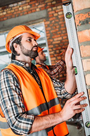 side view of handsome builder using bubble level at construction site while his colleague working on backgroundの写真素材