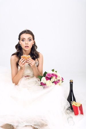 shocked young bride in wedding dress sitting on floor with burger in hand on whiteの写真素材