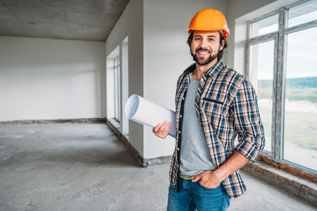handsome smiling architect in plaid shirt and hard hat standing inside of building house with blueprint and looking at cameraの写真素材