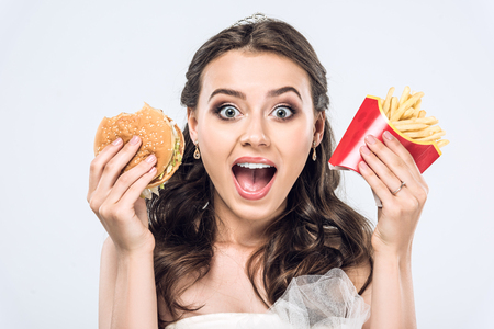 close-up portrait of shocked young bride in wedding dress with burger and french fries looking at camera isolated on whiteの写真素材