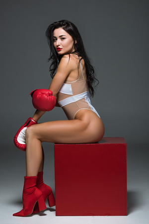 side view of sexy sportive woman in white leotard and boxing gloves sitting on red cube on greyの写真素材