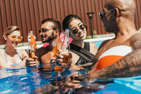 multicultural friends with beach ball and bottles of beer in water in swimming poolの写真素材