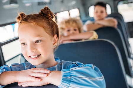 close-up portrait of happy little schoolgirl riding on school bus with classmates behind