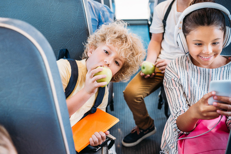 adorable little schoolboy eating apple while riding on school bus with classmates