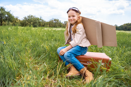 smiling cute kid in pilot costume sitting on retro suitcase in summer fieldの写真素材