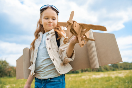 red hair kid in pilot costume holding wooden plane in summer fieldの写真素材