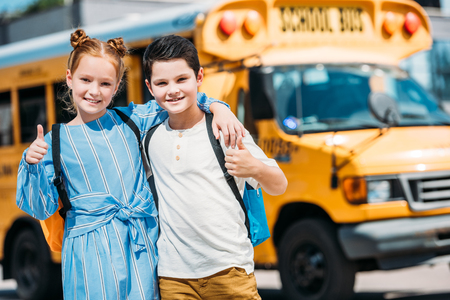 happy little scholars showing thumbs up and looking at camera in front of school bus