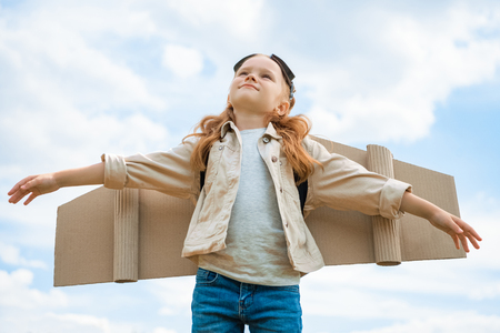 low angle view of child with paper plane wings and protective eyeglasses with outstretched arms against blue cloudy skyの写真素材