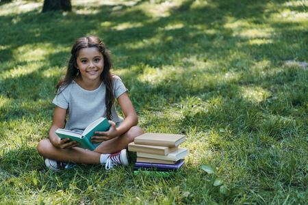 cute happy child sitting in grass with books and smiling at camera in park
