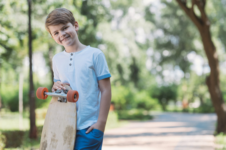 happy kid standing with skateboard and smiling at camera in parkの写真素材