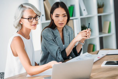cheerful caucasian businesswoman pointing on something at laptop to asian colleague in officeの写真素材