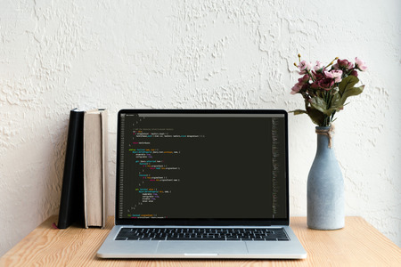 laptop with html code on screen, books and flowers in vase on wooden tableのeditorial素材