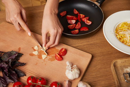 cropped shot of woman cutting garlic and tomatoes for pasta on kitchen tableの写真素材