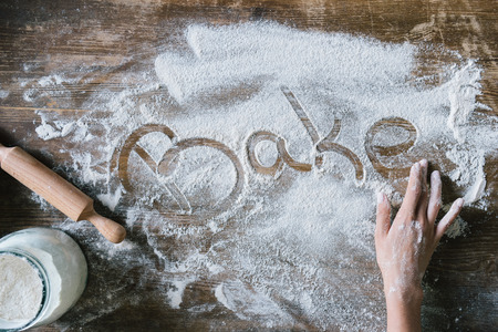 cropped shot of woman writing word BAKE with finger on rustic wooden table covered with flour
