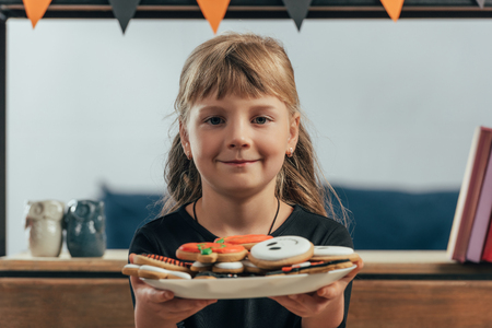 portrait of smiling child showing plate with halloween cookies at homeの写真素材