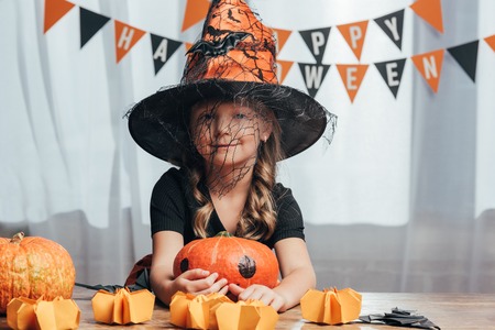 portrait of adorable kid in witch halloween costume at tabletop with pumpkins at homeの写真素材