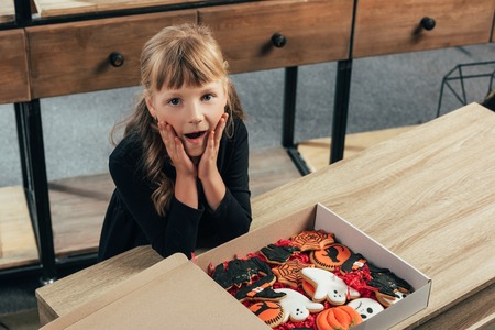 high angle view of shocked kid looking at camera at tabletop with halloween cookies in box at home