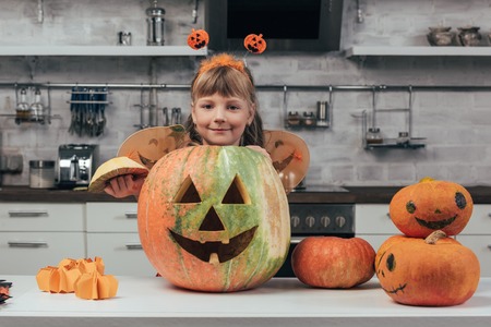 portrait of smiling kid in halloween costume standing at big carved pumpkin on tabletop in kitchen...